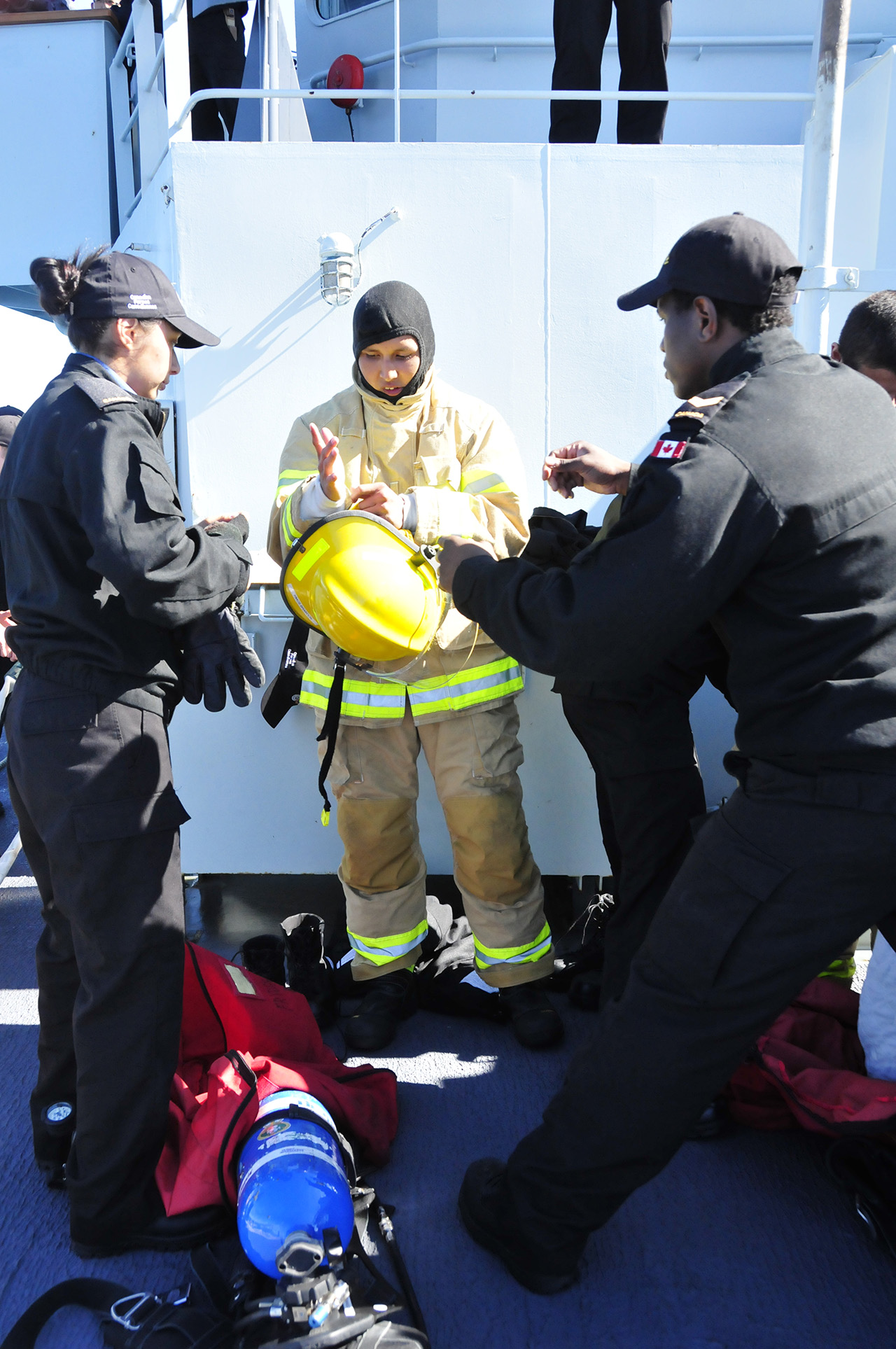 Un participant au programme RAVEN aide un camarade à enfiler l’équipement de lutte contre l’incendie avec l’aide d’un membre d’équipage du NCSM Brandon.