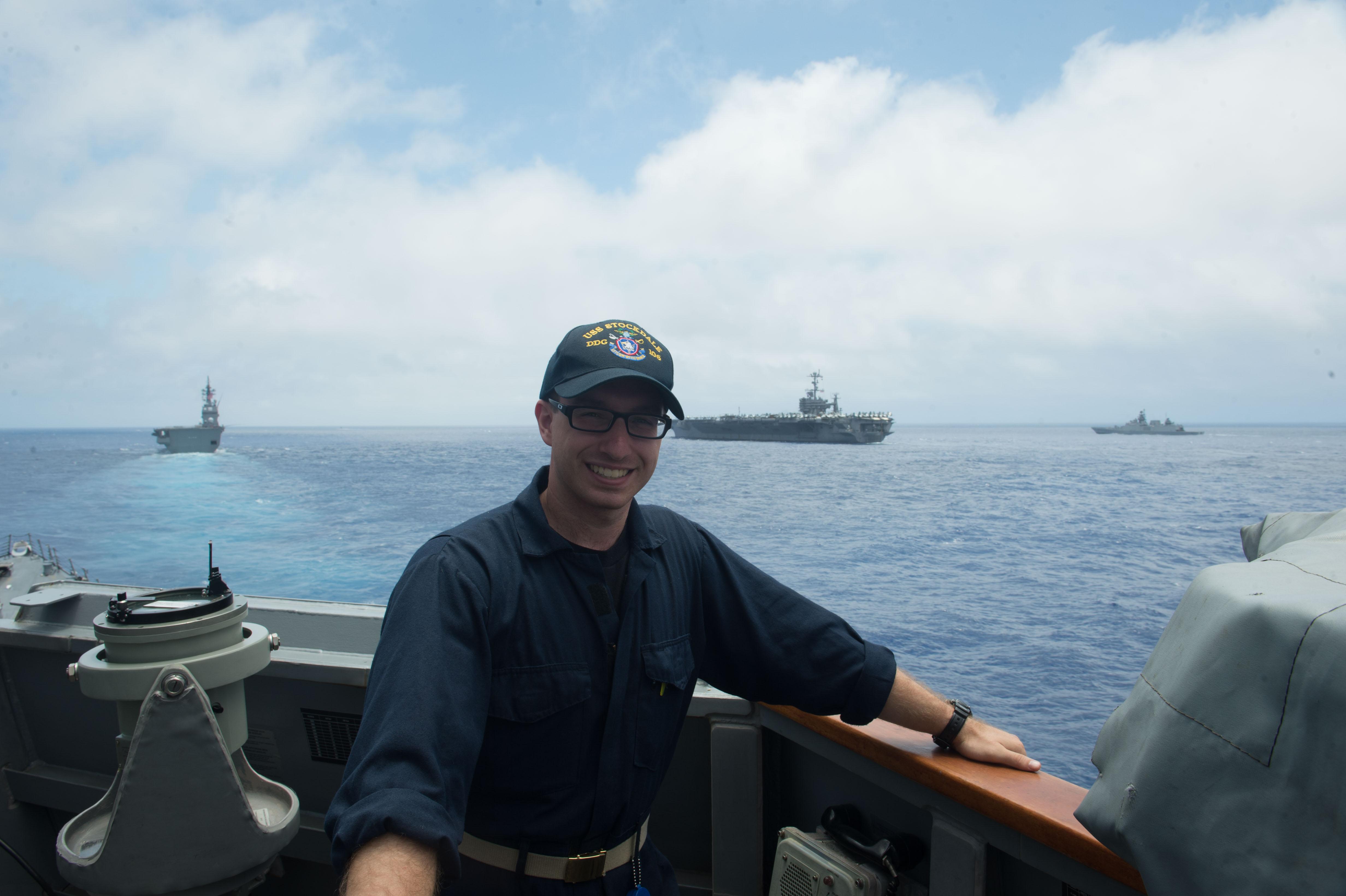 L’aspirant de marine Alek Robaczewski est debout à l’extérieur sur l’aileron de passerelle de l’USS Stockdale, avec trois navires dans la mer des Philippines à l’arrière-plan.
